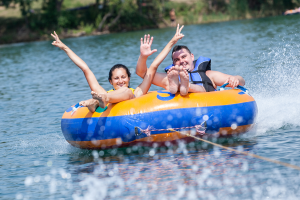 Echuca Moama Boat and Storage Solutions. Photo of a man and a woman in a blue and yellow ring shaped floatation device being pulled by a boat. Both people in the boat are smiling and waving.